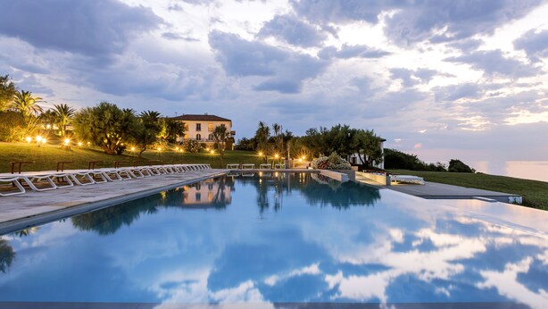 Un área de piscina tranquila, con un reflejo nítido del cielo, rodeada de exuberante vegetación y un edificio bien iluminado al fondo.