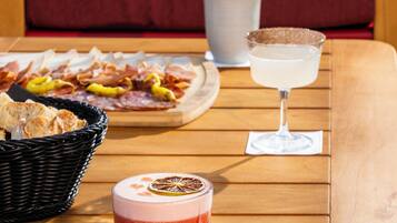 A wooden table with a red beverage, a glass of white drink, a basket of bread, and a platter of meats.