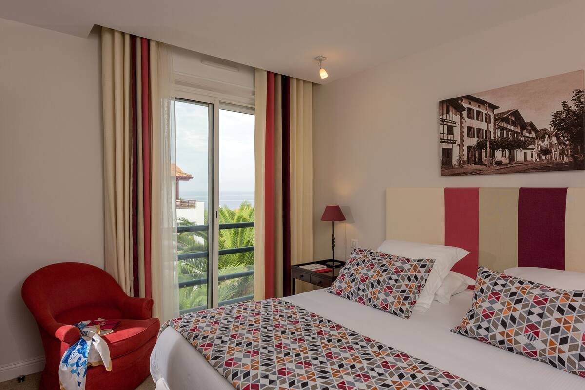 Un cuarto de hotel con cama, un sillón rojo, una mesita de noche, una lámpara y vistas a un balcón con palmeras.
