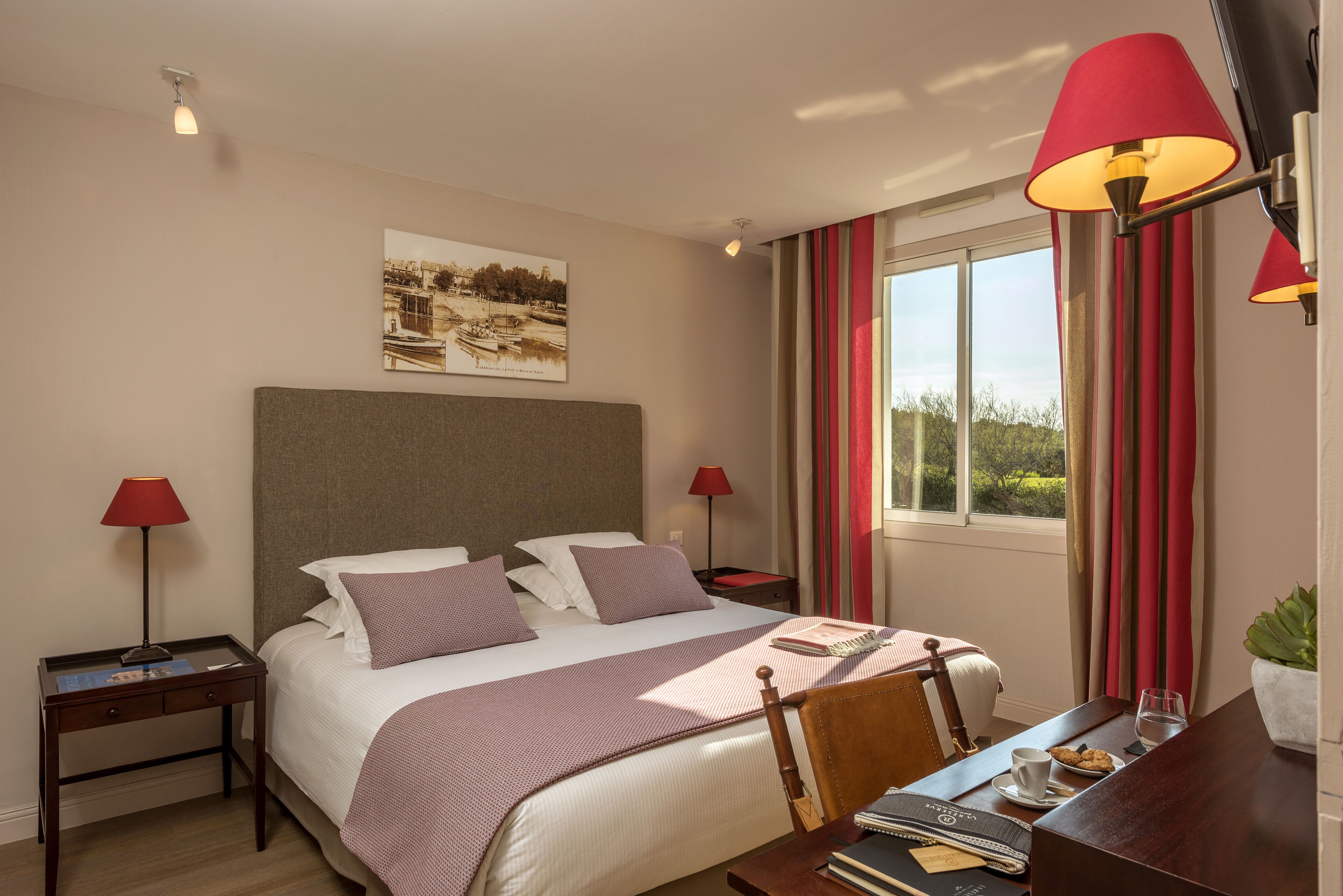 Una habitación de hotel con cama, dos mesitas de noche, un escritorio con silla, una ventana con cortinas y una lámpara de pared.