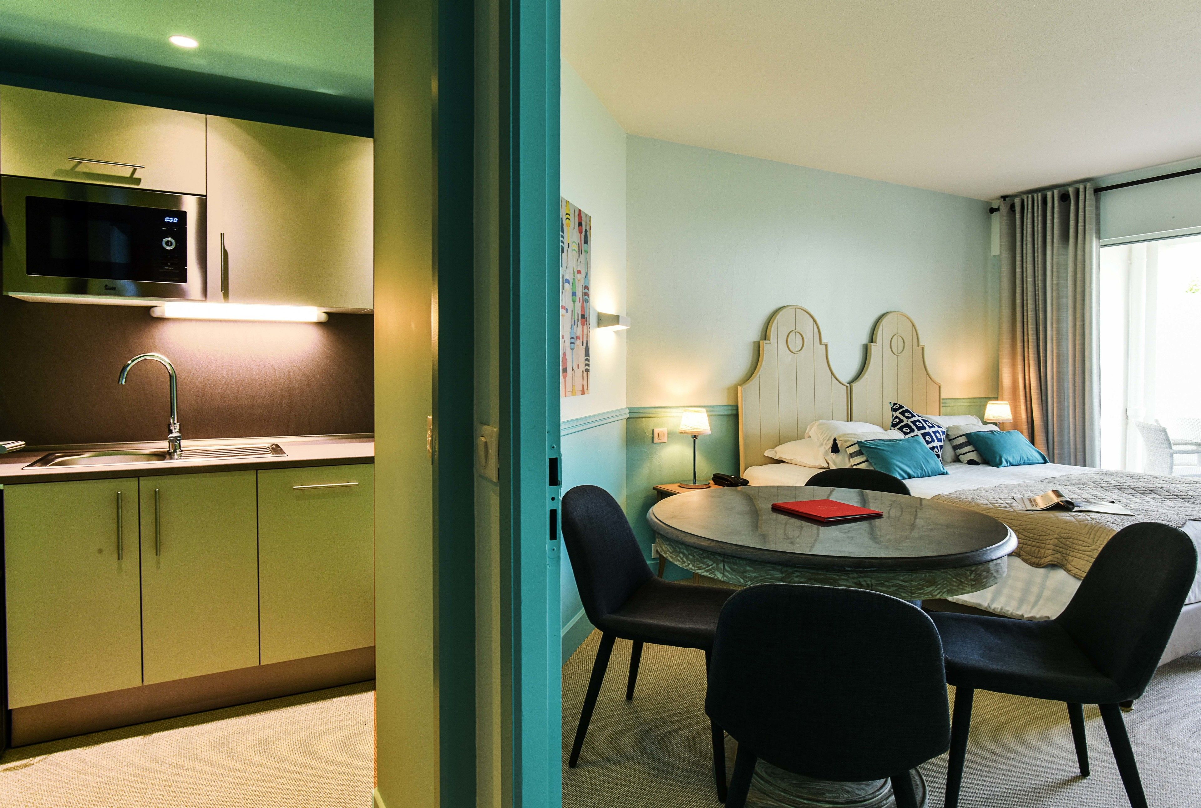 A hotel room with a kitchenette, a bed with a headboard, a round table with chairs, and a nightstand.