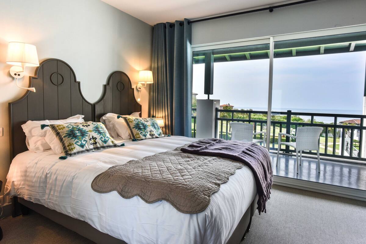 Habitación de hotel con una cama grande, dos lámparas de pared, un balcón con vistas y una ventana con cortinas.