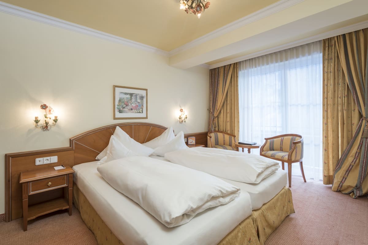 Une chambre d’hôtel avec un lit double, des tables de chevet, une petite table de nuit, une chaise, une petite table, un luminaire fixé au mur et une fenêtre avec des rideaux.