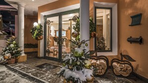 Entrada de un hotel con un árbol de Navidad cubierto de nieve, un espejo grande que refleja el interior, y una exhibición de troncos de madera en el exterior.