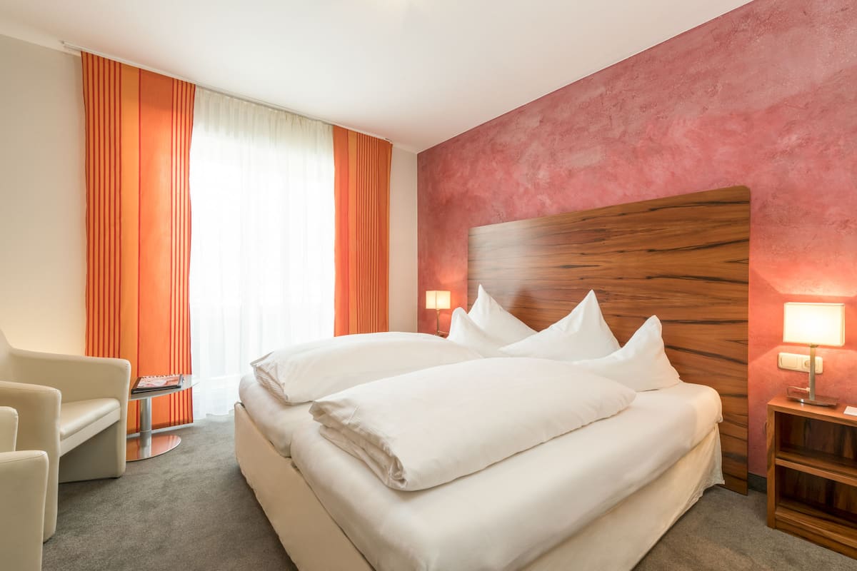 Une chambre d’hôtel avec un grand lit, une chaise, une table de chevet avec une lampe et une fenêtre avec des rideaux.