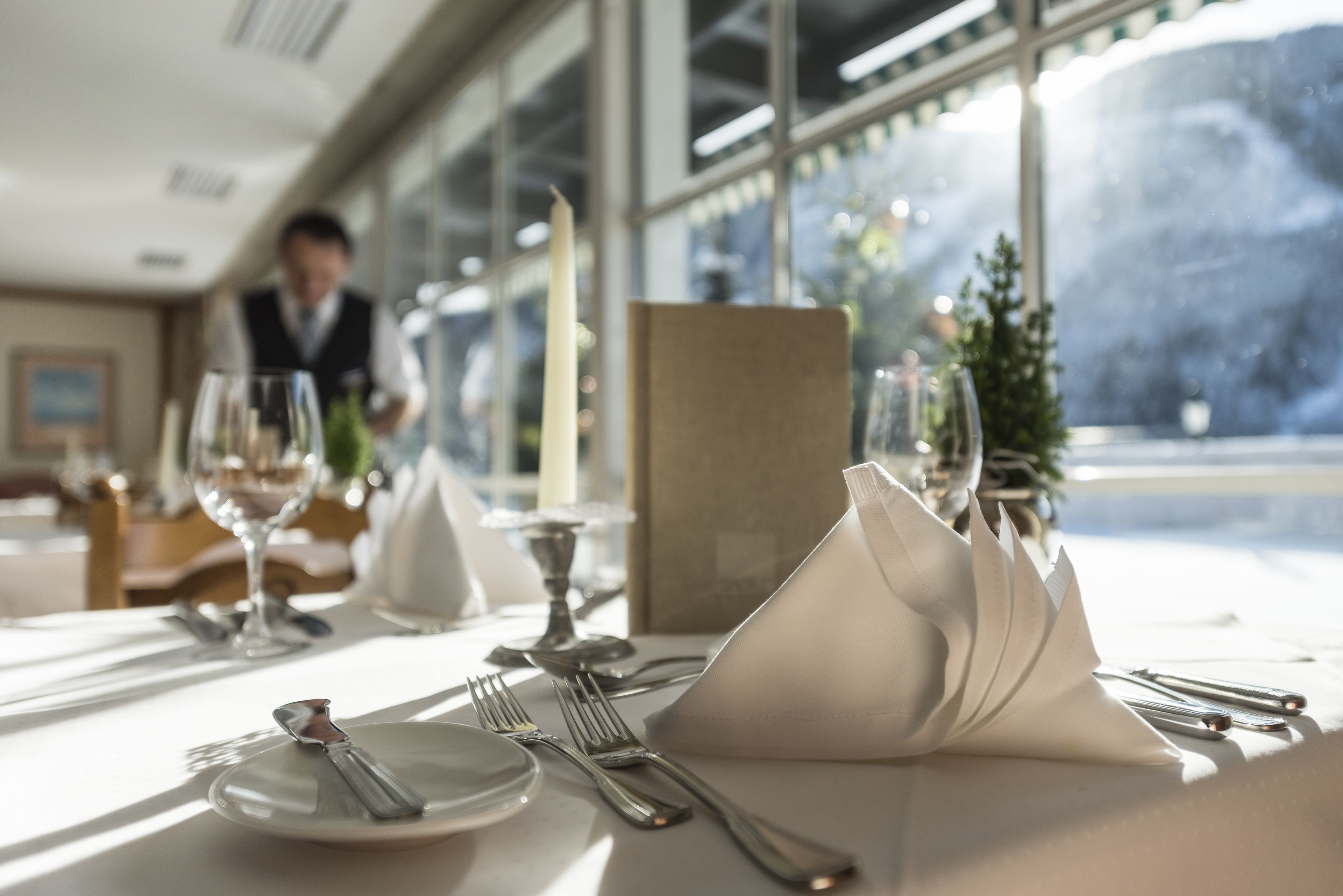 A well-set dining table with a candle, a folded napkin, and cutlery, with a view of a snowy landscape through large windows.
