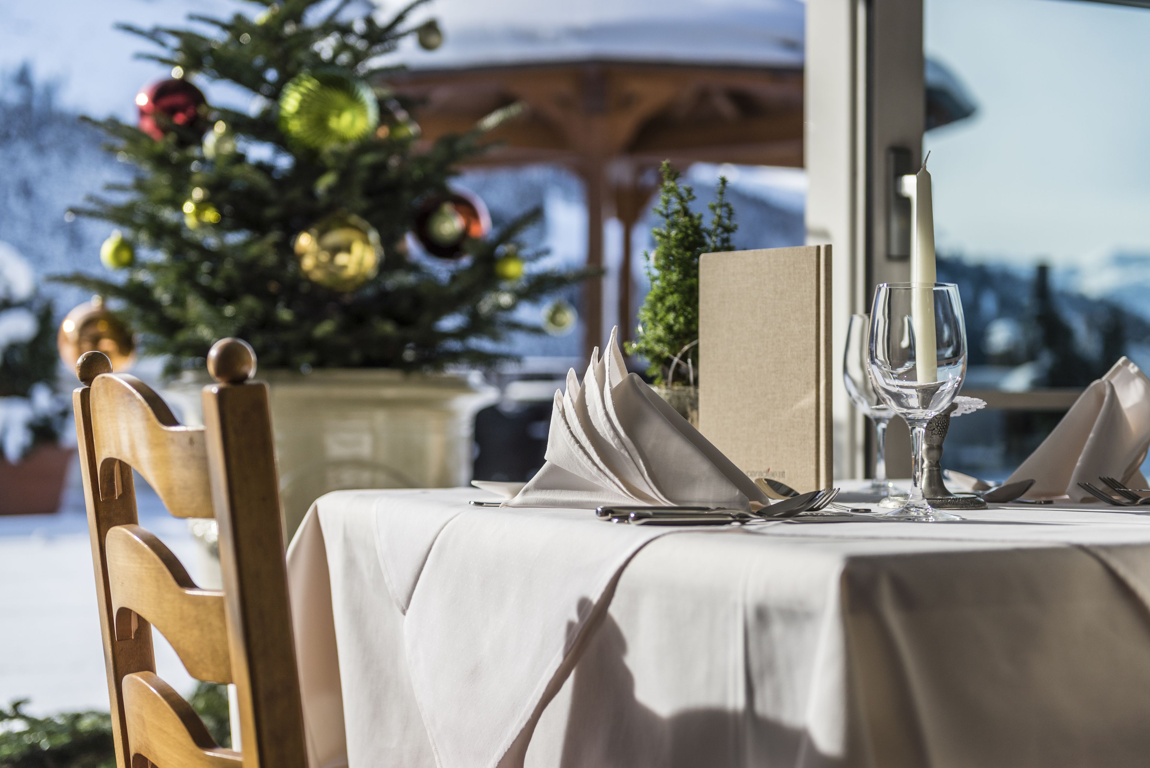 A dining table set with a white tablecloth, a candle, and cutlery, with a Christmas tree in the background.