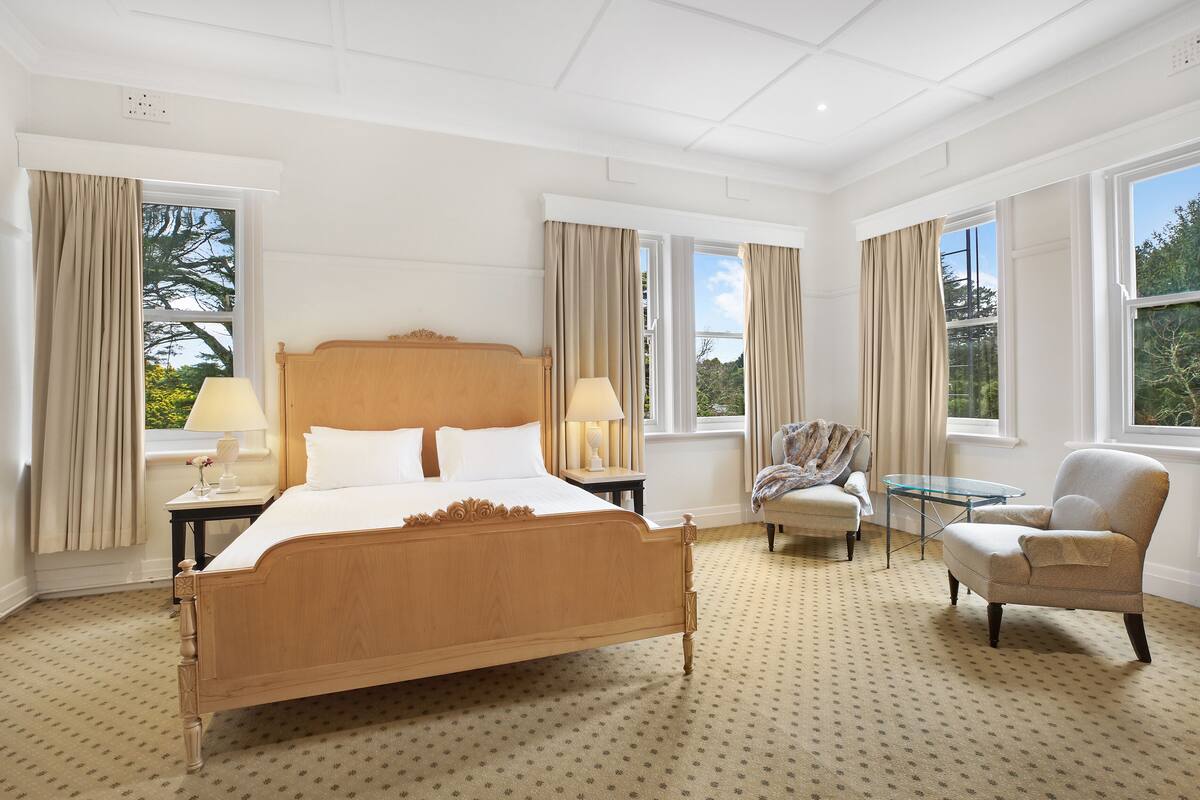 Quarto de hotel com uma cama grande, duas mesas de cabeceira com abajures, uma cadeira e uma pequena mesa.