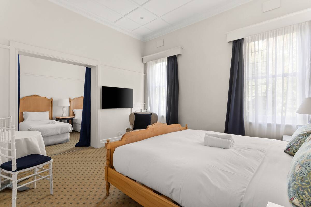 Quarto de hotel com uma cama grande, uma mesa com cadeira, uma TV fixada na parede e uma janela com cortinas.