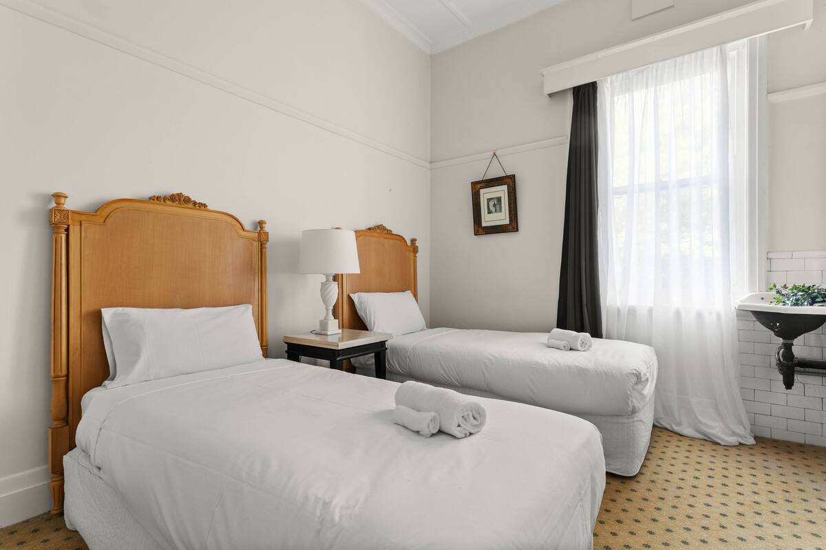 Quarto de hotel com duas camas de solteiro, mesa de cabeceira com abajur, pequena moldura de quadro, janela com cortinas translúcidas e pia com espelho.
