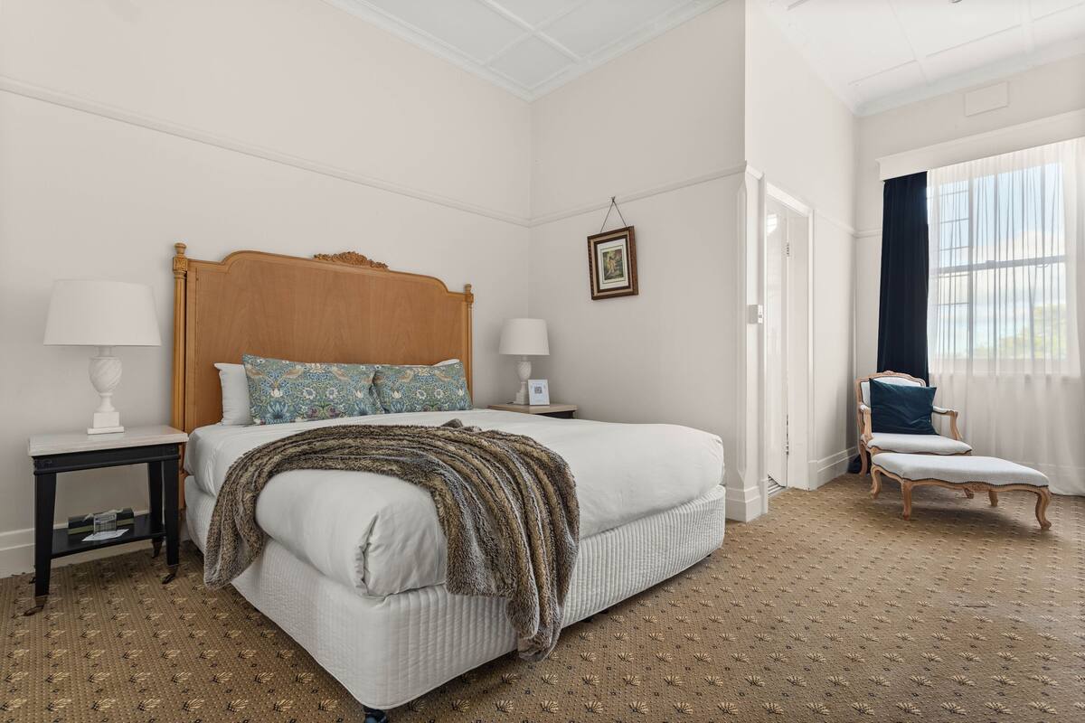 Quarto com uma cama grande, duas mesas de cabeceira, uma cadeira e uma janela com cortinas.