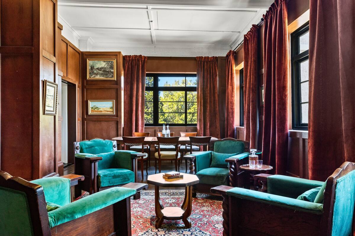 Quarto com paredes revestidas de madeira, cadeiras estofadas em verde, uma mesa redonda com um livro e uma janela grande com cortinas.