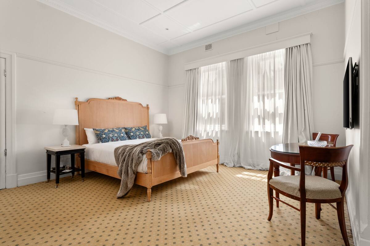 Quarto de hotel com cama de madeira, uma mesa de jantar redonda, uma cadeira e televisão na parede.
