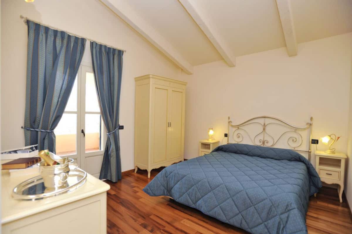 A bedroom with a bed, a nightstand, a wardrobe, a window with curtains, and a bedside table with a lamp.