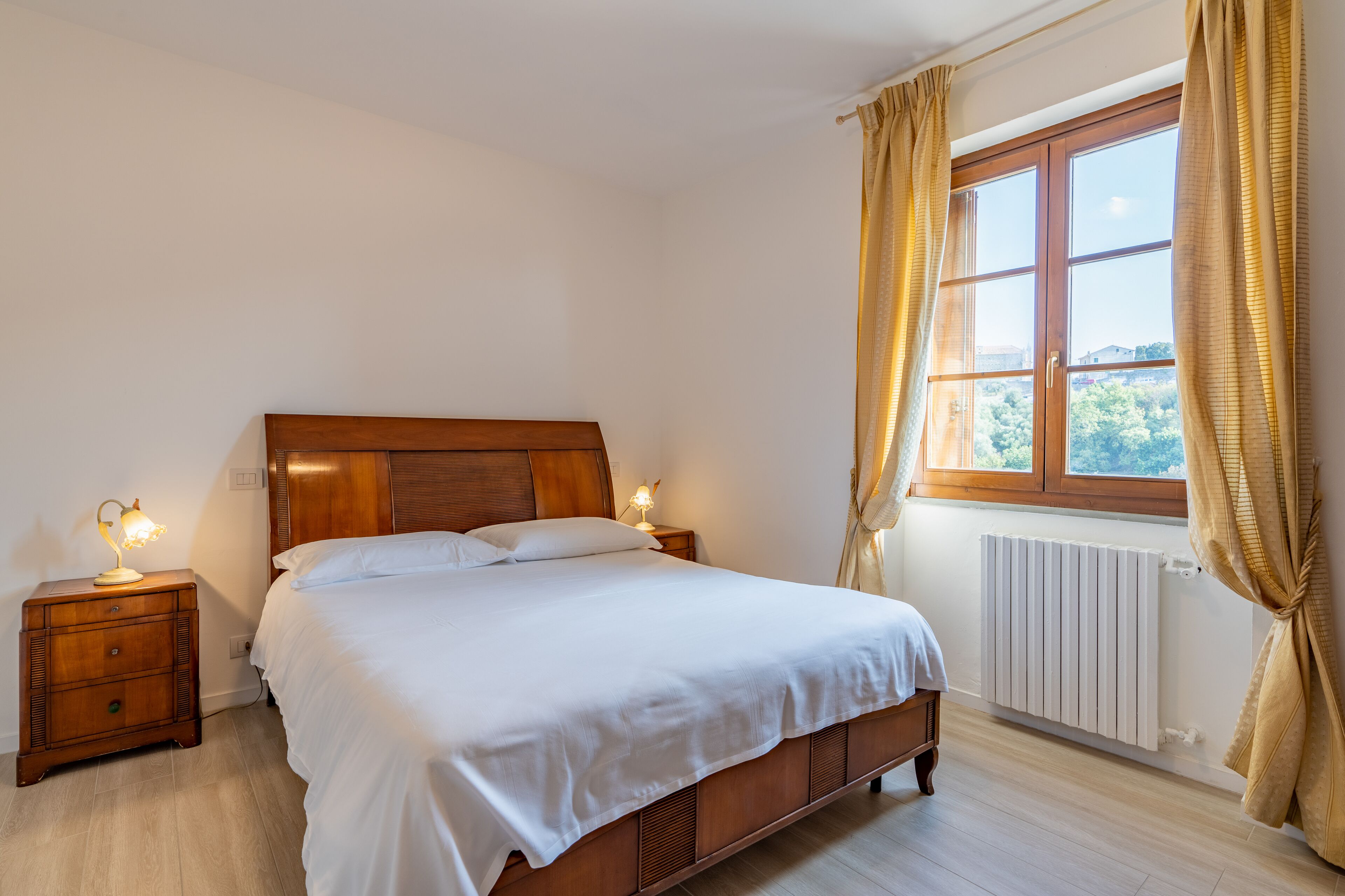 A neatly made bed with a wooden headboard, two bedside tables with lamps, a window with curtains, and a radiator.