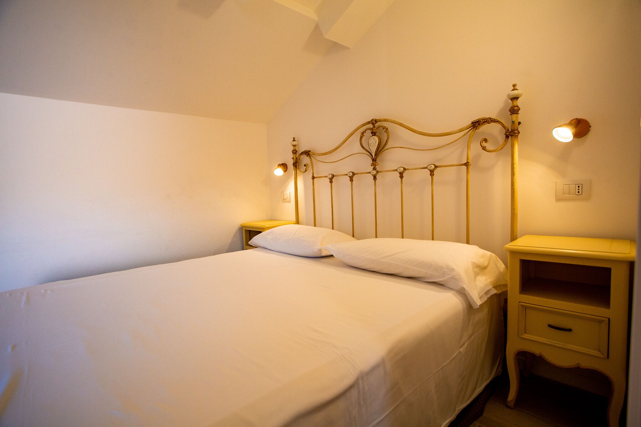 A single bed with a metal headboard, two bedside tables with lamps, and a small drawer unit.