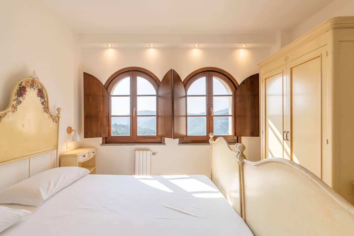 A bedroom with a bed, two windows with wooden shutters, a bedside table with a lamp, and a wardrobe.
