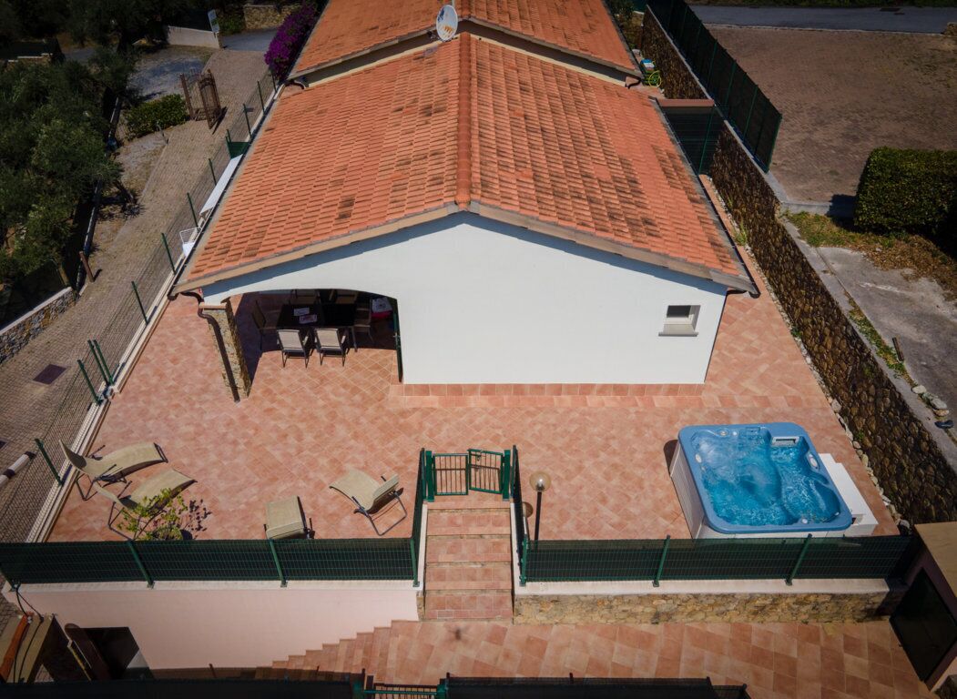An aerial view of a building with a tiled roof, a covered patio area with chairs, a hot tub, and a staircase leading to an upper level.