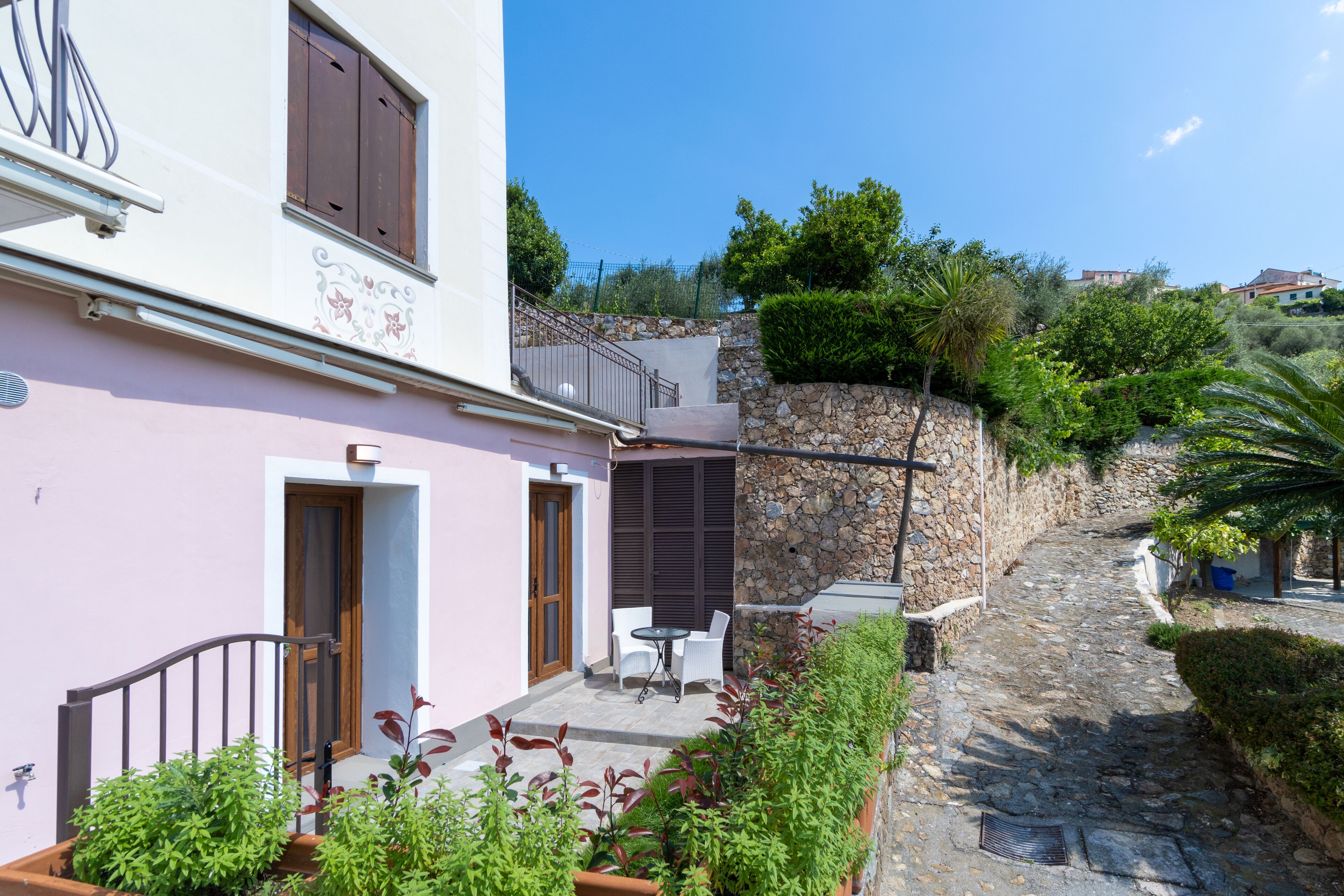 Edificio de paredes de piedra con fachada rosa, un pequeño patio con mesa y sillas, y un sendero de piedra que lleva a una puerta.