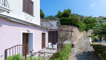 A stone-walled building with a pink facade, a small patio with a table and chairs, and a stone pathway leading to a gate.