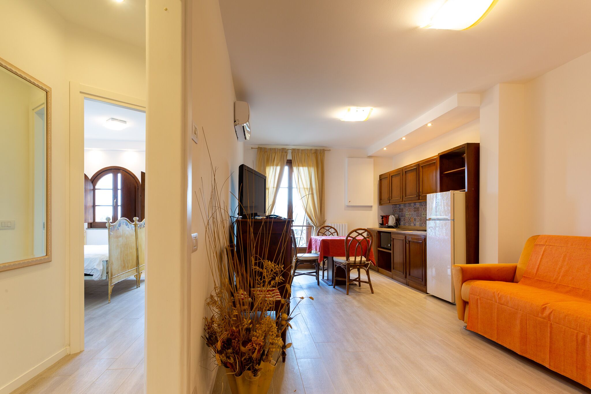 Una sala de estar con sofá, una mesa de comedor con sillas, una zona de cocina con armarios y refrigerador, y un dormitorio con cama y espejo.
