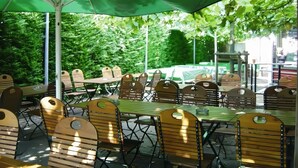 An outdoor seating area with tables and chairs, covered by a green umbrella, surrounded by lush greenery. - Hotel Brößler (Stockstadt am Main)