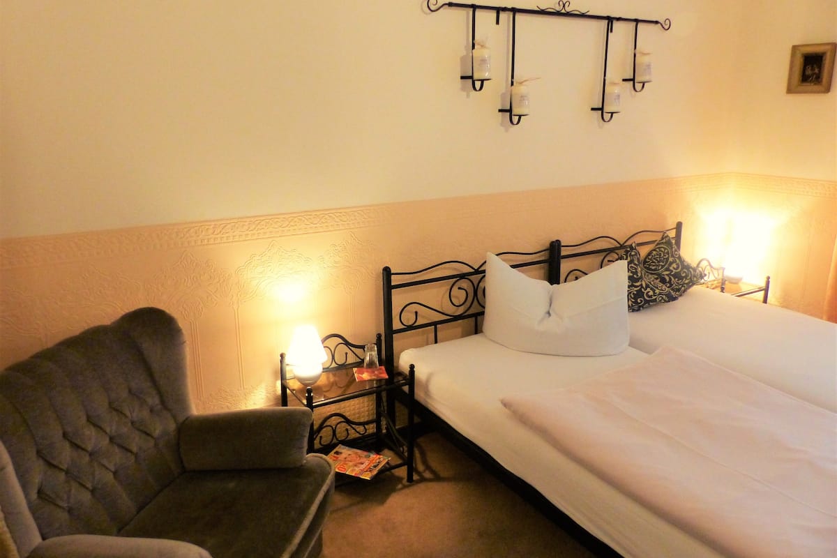 Une chambre d’hôtel avec un lit, une chaise, une table de chevet et un luminaire fixé au mur.