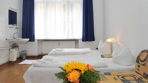 Quarto de hotel com duas camas, banheiro, janela com cortinas, mesa de cabeceira com abajur e um vaso de flores.