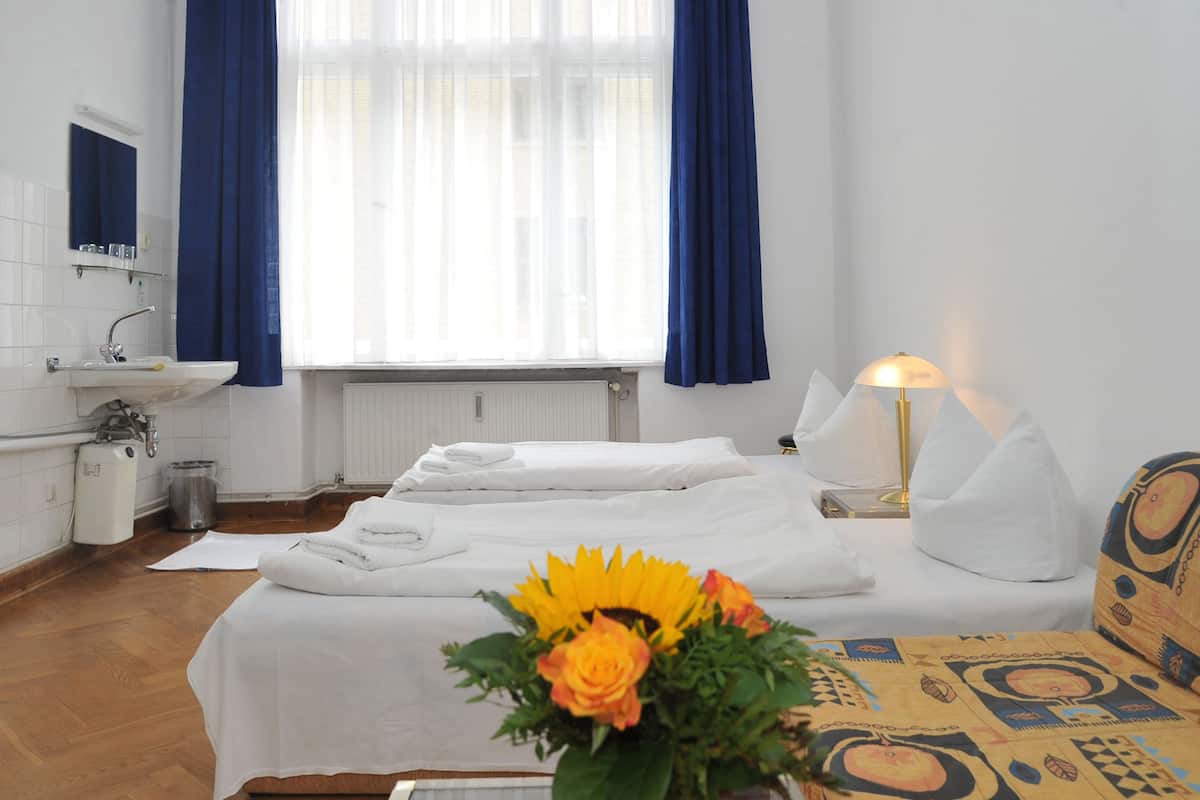 Une chambre d’hôtel avec deux lits, une salle de bain, une fenêtre avec des rideaux, une table de chevet avec une lampe et un vase à fleurs.