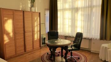 Ein Zimmer mit einem runden Tisch, zwei Stühlen, einem Kleiderschrank und einem Fenster mit durchsichtigen Vorhängen.