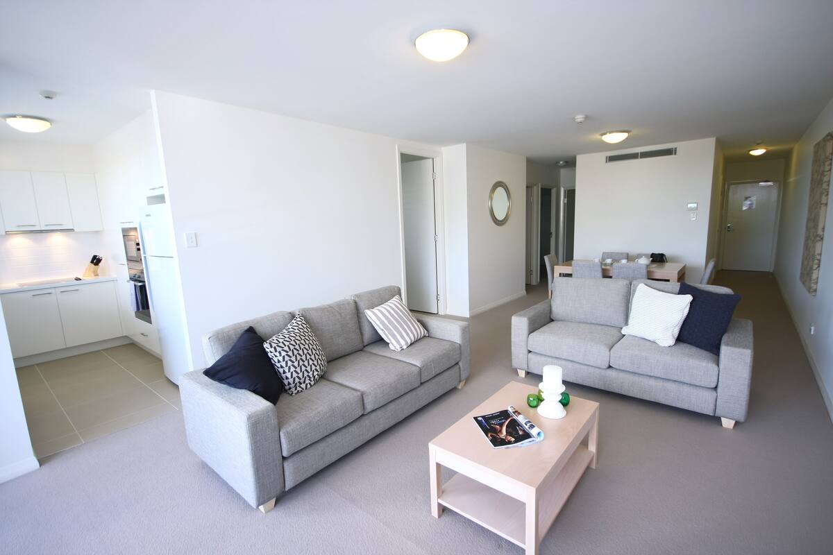 A modern living room with two grey sofas, a coffee table, and a kitchen in the background.