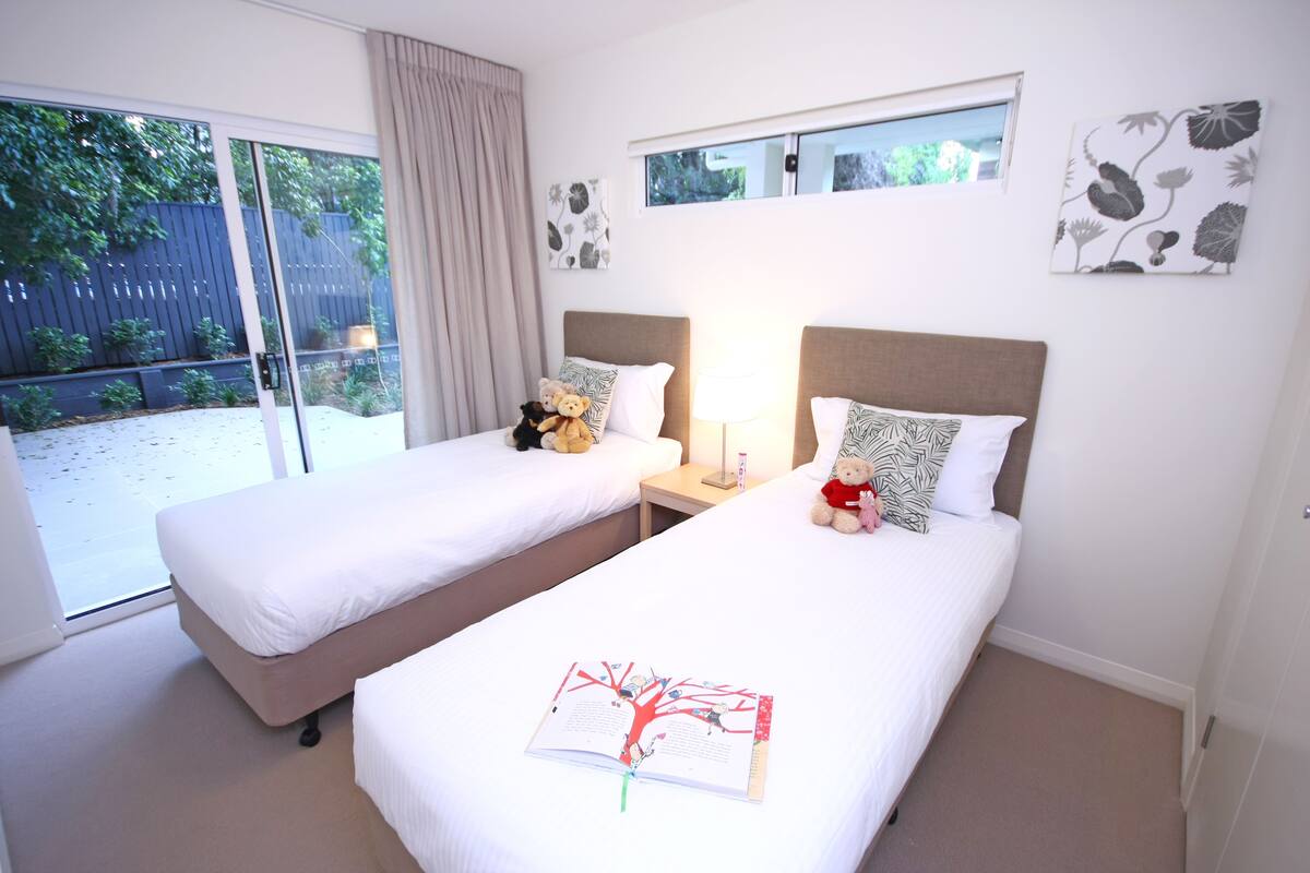 A hotel room with two single beds, a large window with a view of a backyard, and a small table with a lamp.
