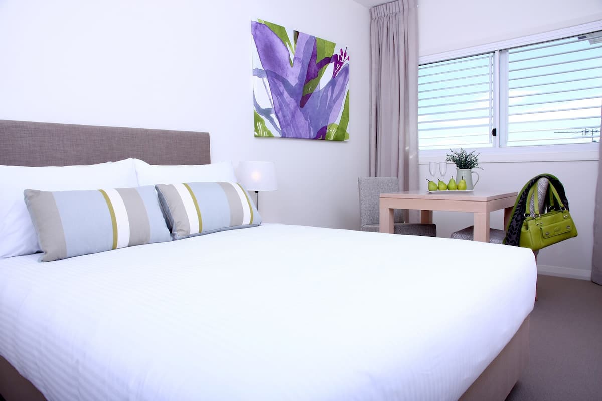 A neatly made bed with striped pillows, a wall-mounted artwork, a bedside table with a lamp, and a window with blinds.