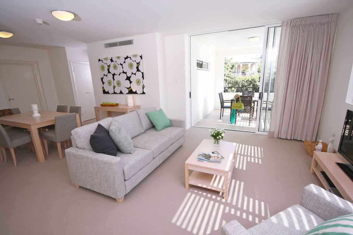 A modern living room with a grey sofa, a coffee table, and a TV stand. There is a dining area with a table and chairs, and a balcony with a view of the outside.