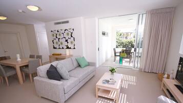 A modern living room with a grey sofa, a coffee table, and a TV stand. There is a dining area with a table and chairs, and a balcony with a view of the outside.