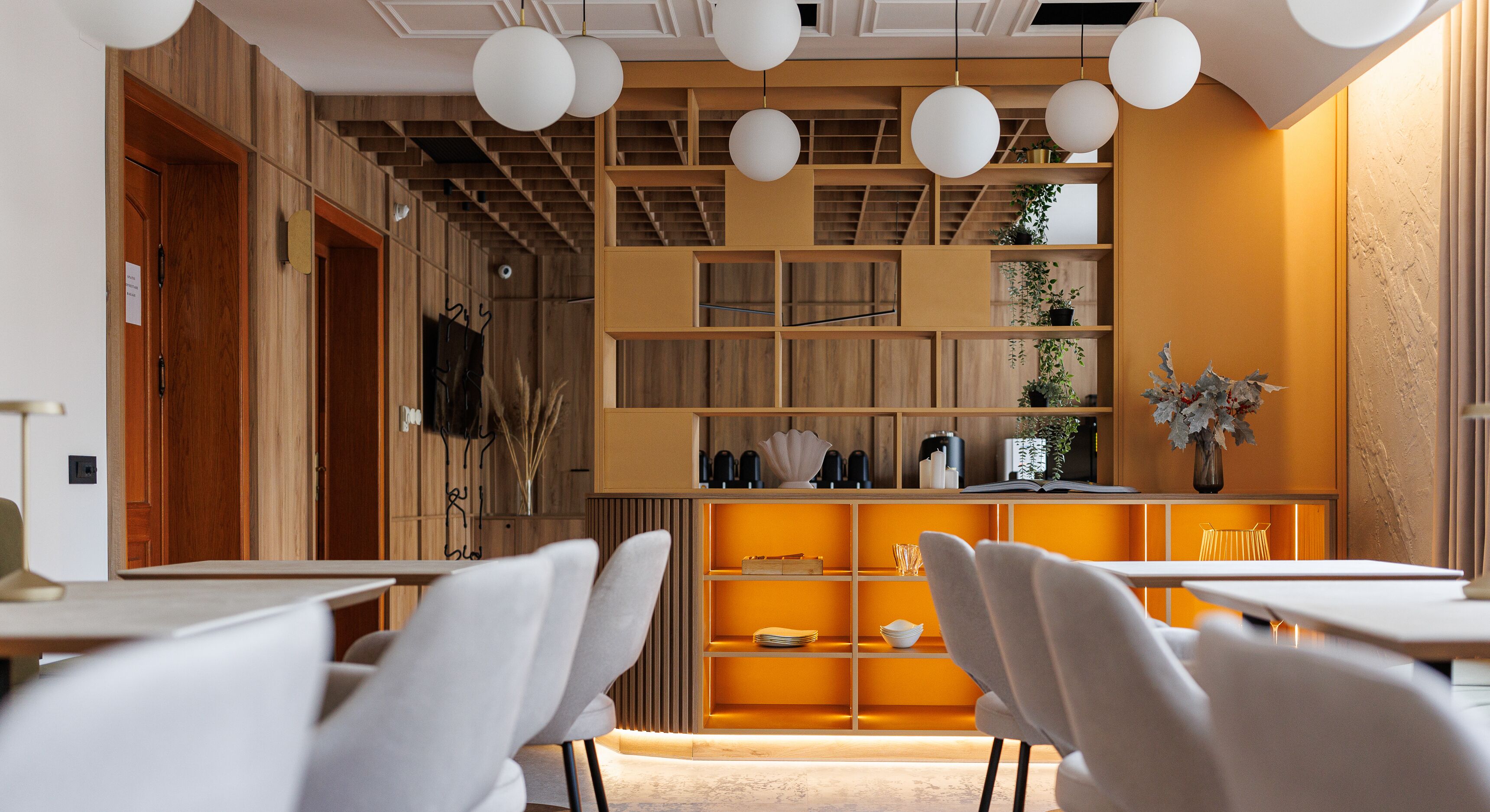Un intérieur moderne avec des murs lambrés en bois, une bibliothèque centrale agrémentée de plantes et des chaises blanches disposées autour d’une table.