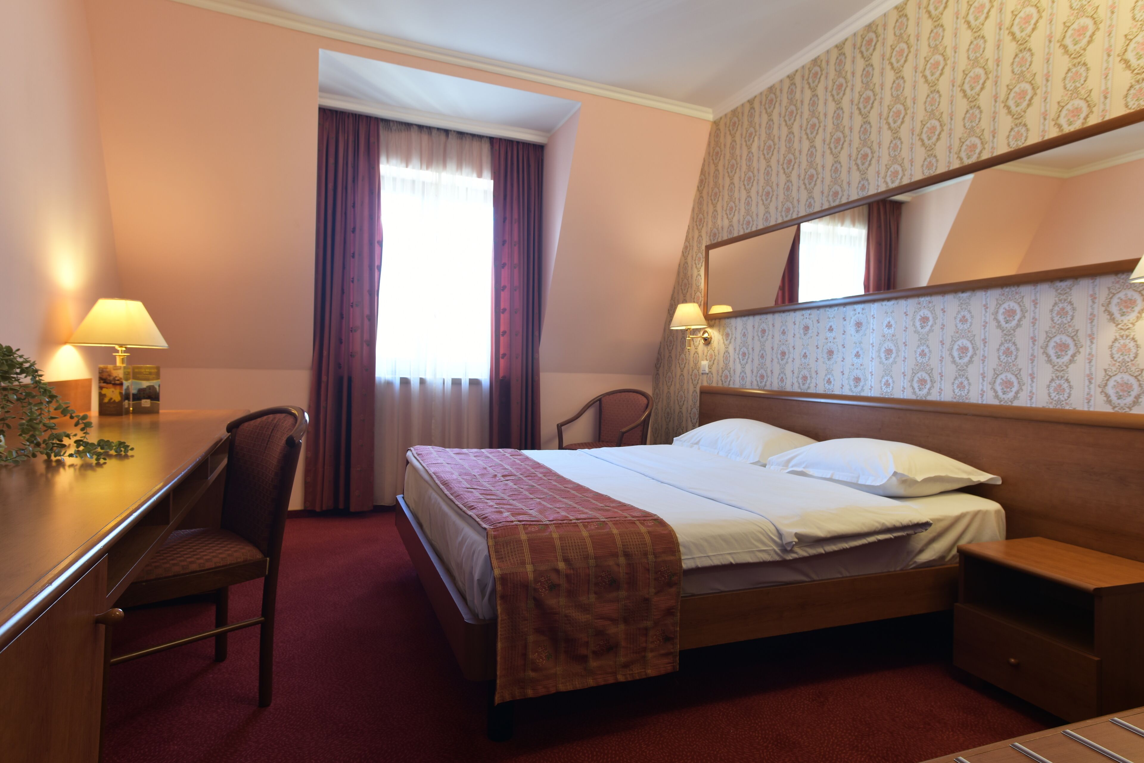 A hotel room with a bed, a desk, a chair, a nightstand, and a mirror.