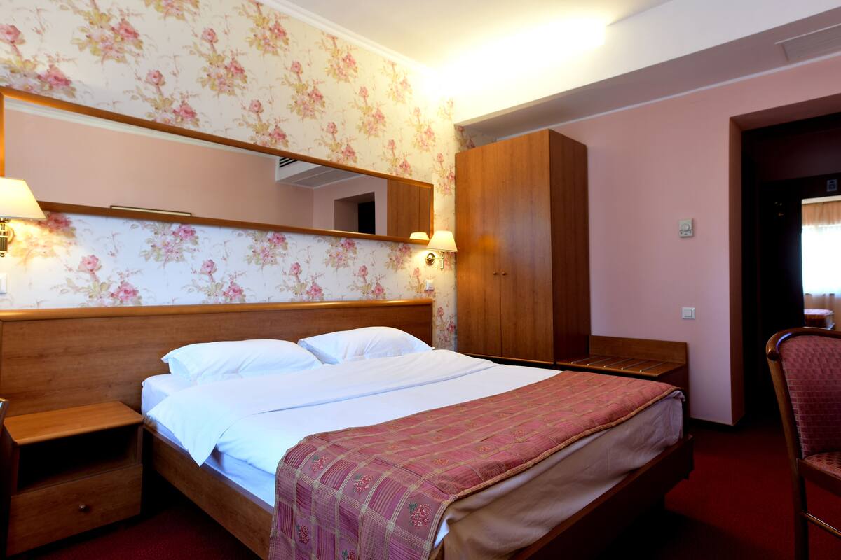 A hotel room with a wooden bed, a nightstand, a wardrobe, and a mirror on the wall.