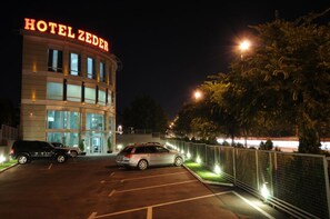 A hotel with a prominent sign, a parking lot with two cars, and a fenced area with trees. - Garni Hotel Zeder (Belgrade)