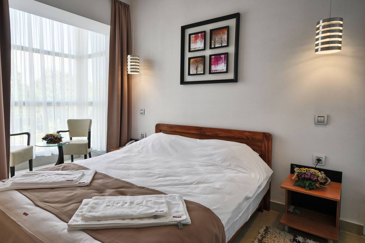 A hotel room with a large bed, a bedside table with a vase of flowers, a chair, and a window with sheer curtains.