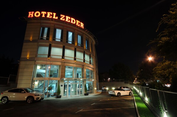 A hotel with a prominent sign, a parking lot with several cars, and a well-lit entrance. - Garni Hotel Zeder (Belgrade)