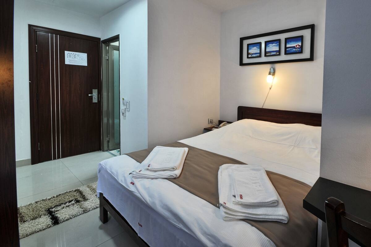 A hotel room with a bed, a desk, a chair, a nightstand, a lamp, and a door.