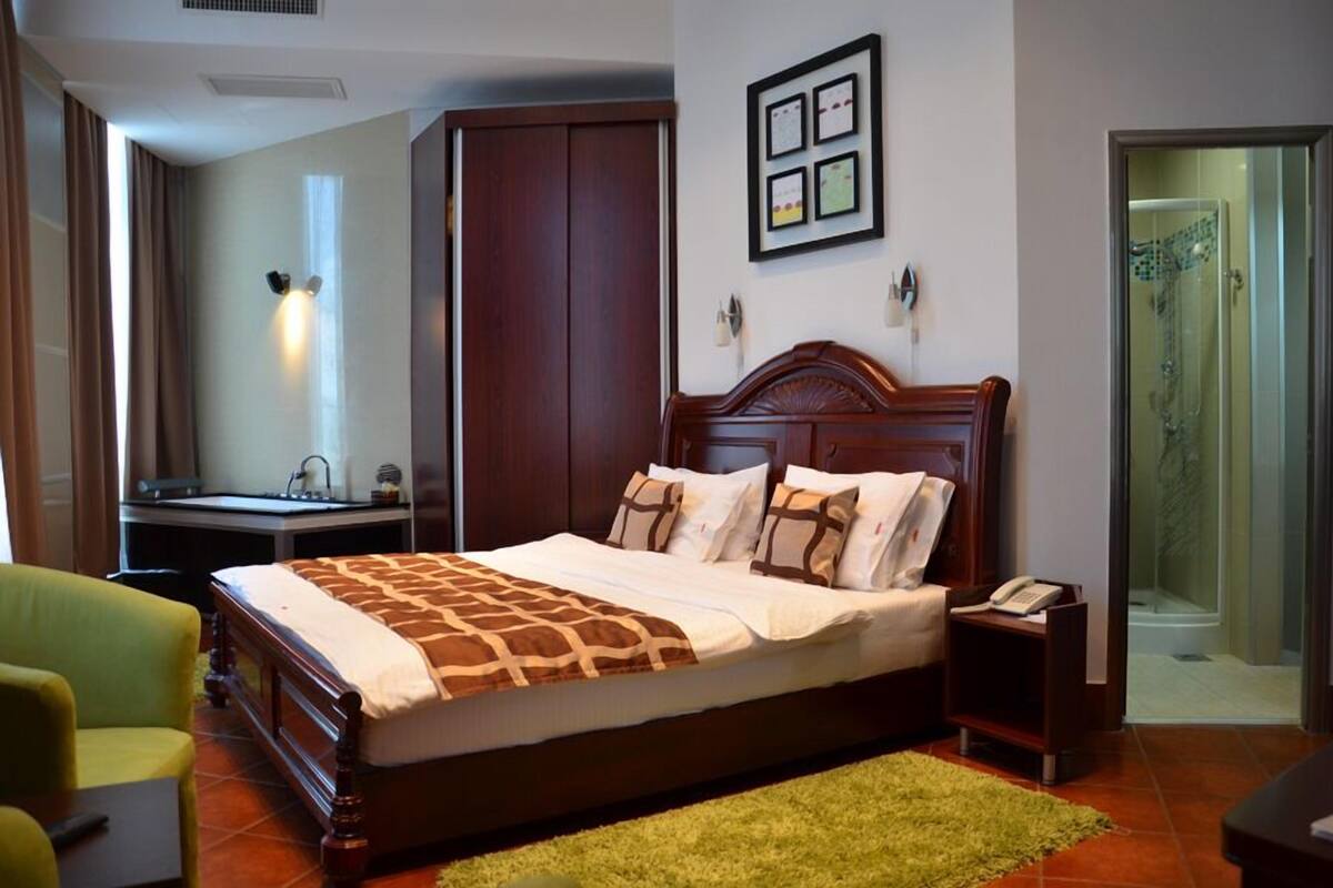 A hotel room with a large bed, a desk with a chair, a green armchair, a wooden wardrobe, and a shower area.