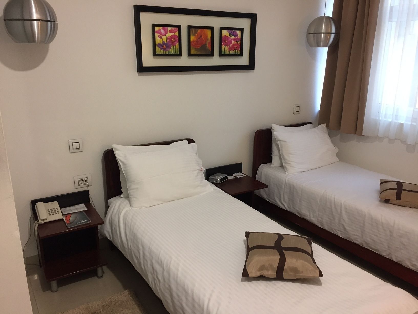 Una habitación de hotel con dos camas individuales, una mesita de noche con un teléfono y un control remoto, una obra de arte floral enmarcada sobre las camas, y una ventana con cortinas.