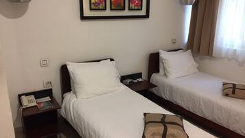 Una habitación de hotel con dos camas individuales, una mesita de noche con un teléfono y un control remoto, una obra de arte floral enmarcada sobre las camas, y una ventana con cortinas.