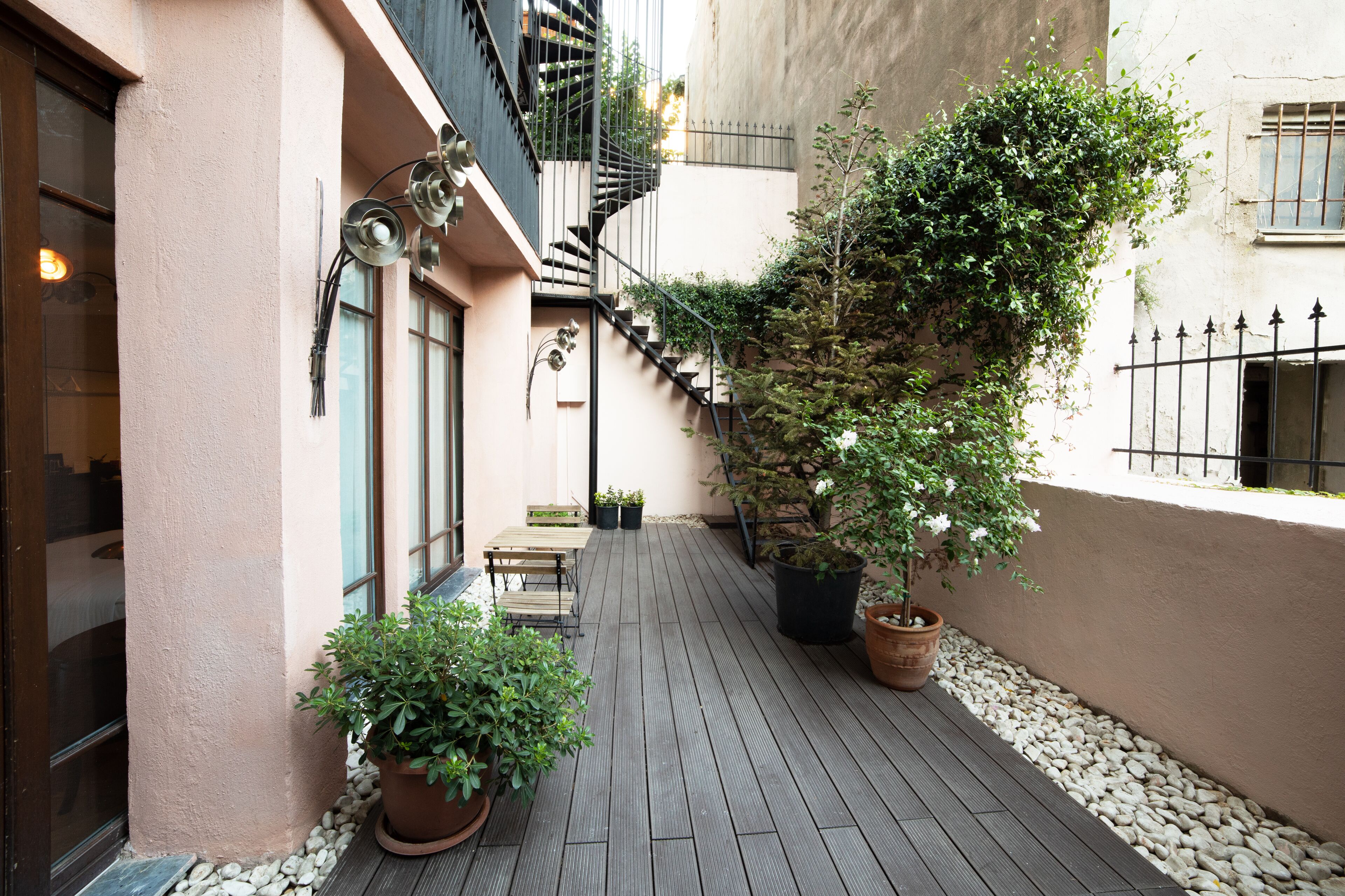 Un étroit couloir extérieur doté d’un plancher en bois, de plantes en pot et d’un escalier menant à un niveau supérieur.