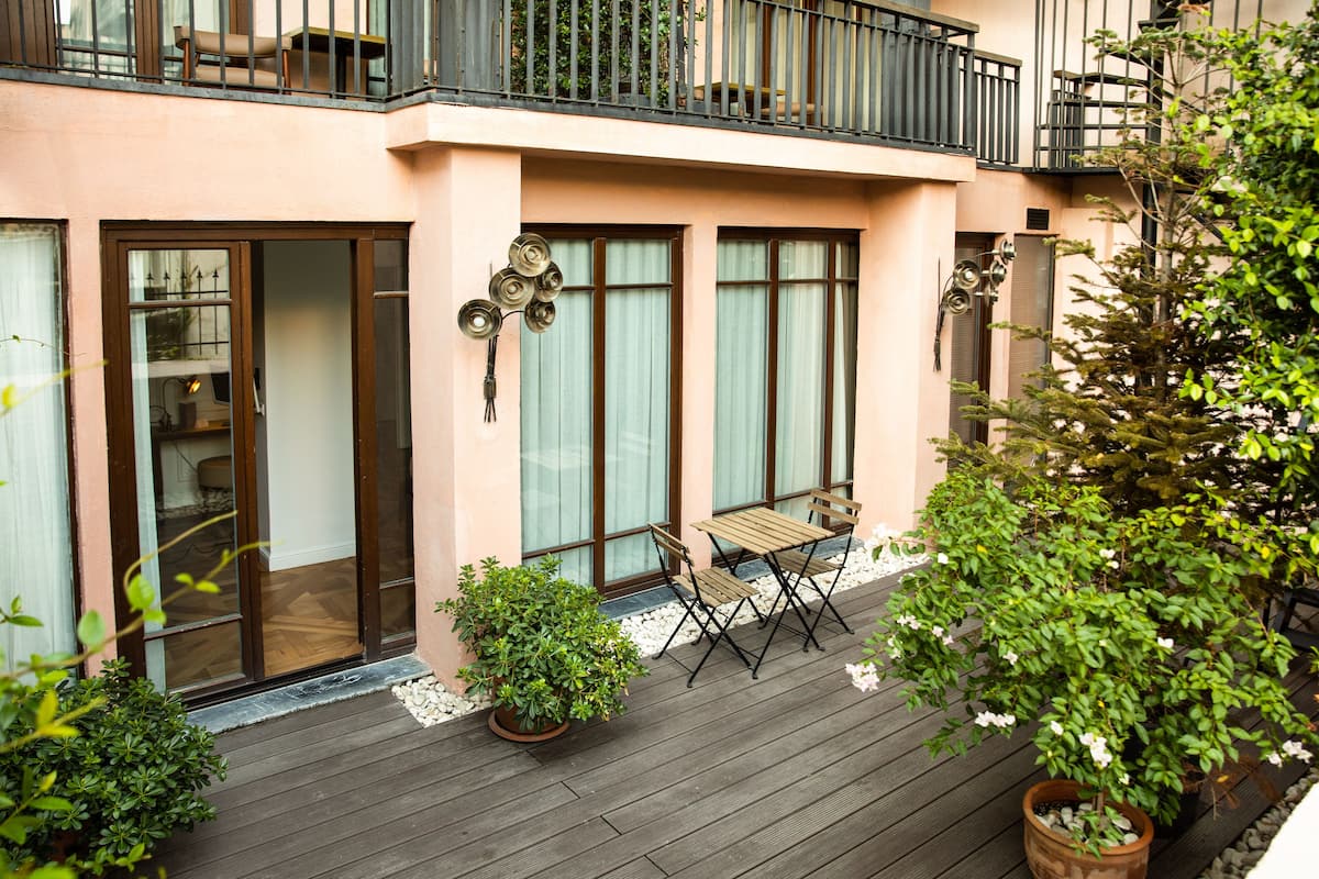 A balcony with a wooden table and chairs, potted plants, and a view of a building with large windows and a balcony.
