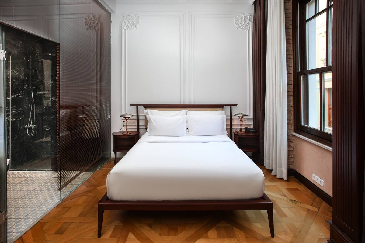 A large bed with a wooden frame and white linens, positioned in a room with wooden flooring, a glass shower enclosure, and a large window with curtains.