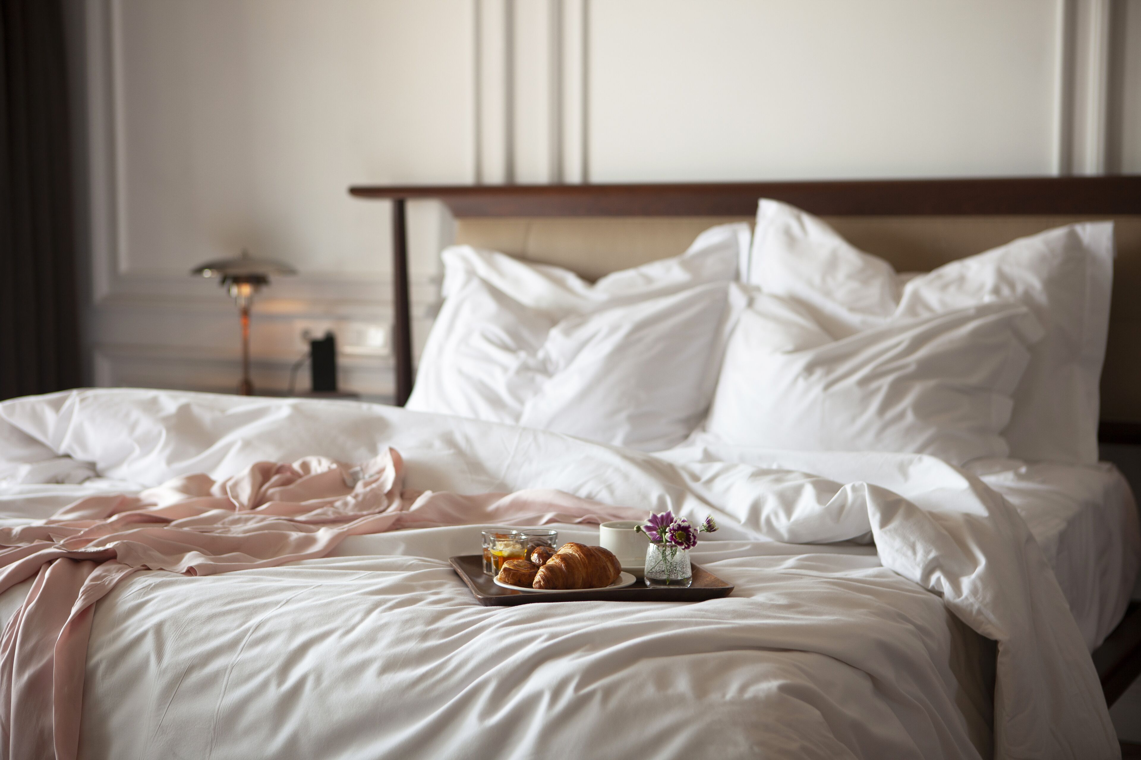 A neatly made bed with white linens, a tray with croissants and a cup of coffee, and a vase with flowers.