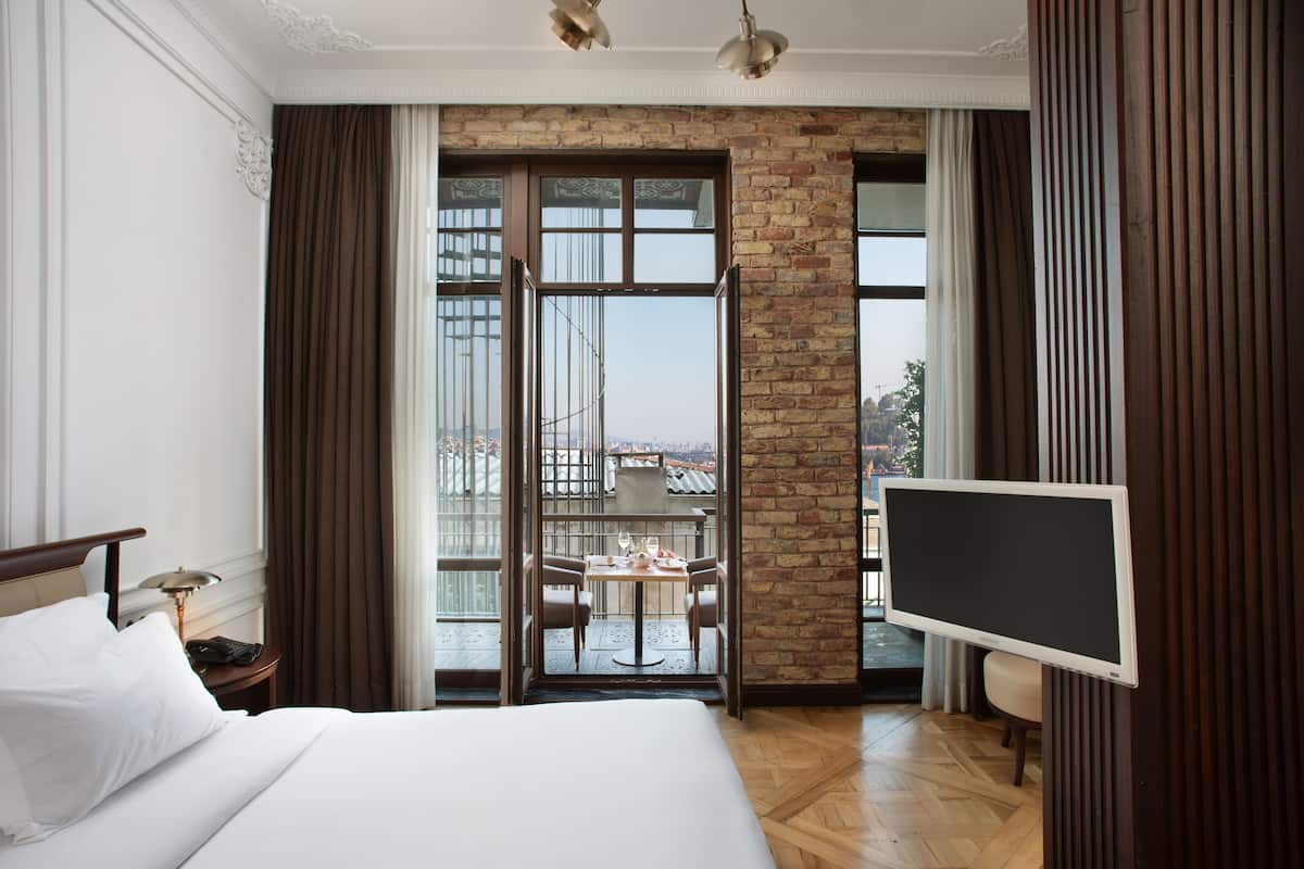 A hotel room with a large bed, a balcony with a table and chairs, a TV, and a view of the city.